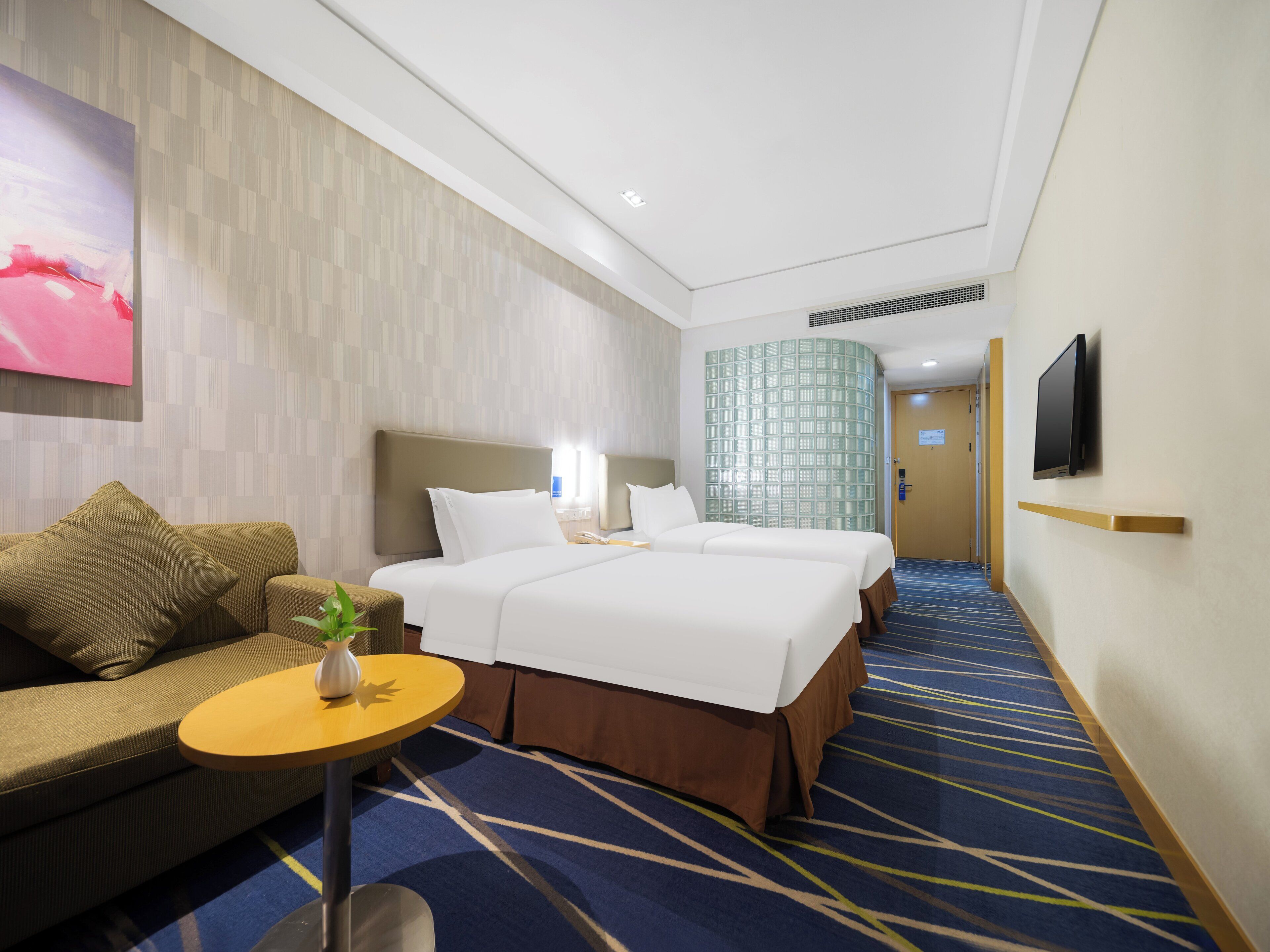 A hotel room with two beds, a sofa, a small table with a plant, a TV mounted on the wall, and a patterned carpet.