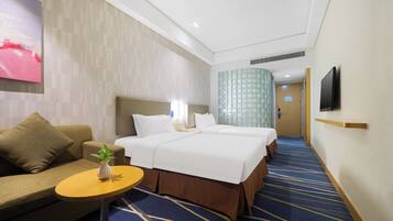 A hotel room with two beds, a sofa, a small table with a plant, a TV mounted on the wall, and a patterned carpet.