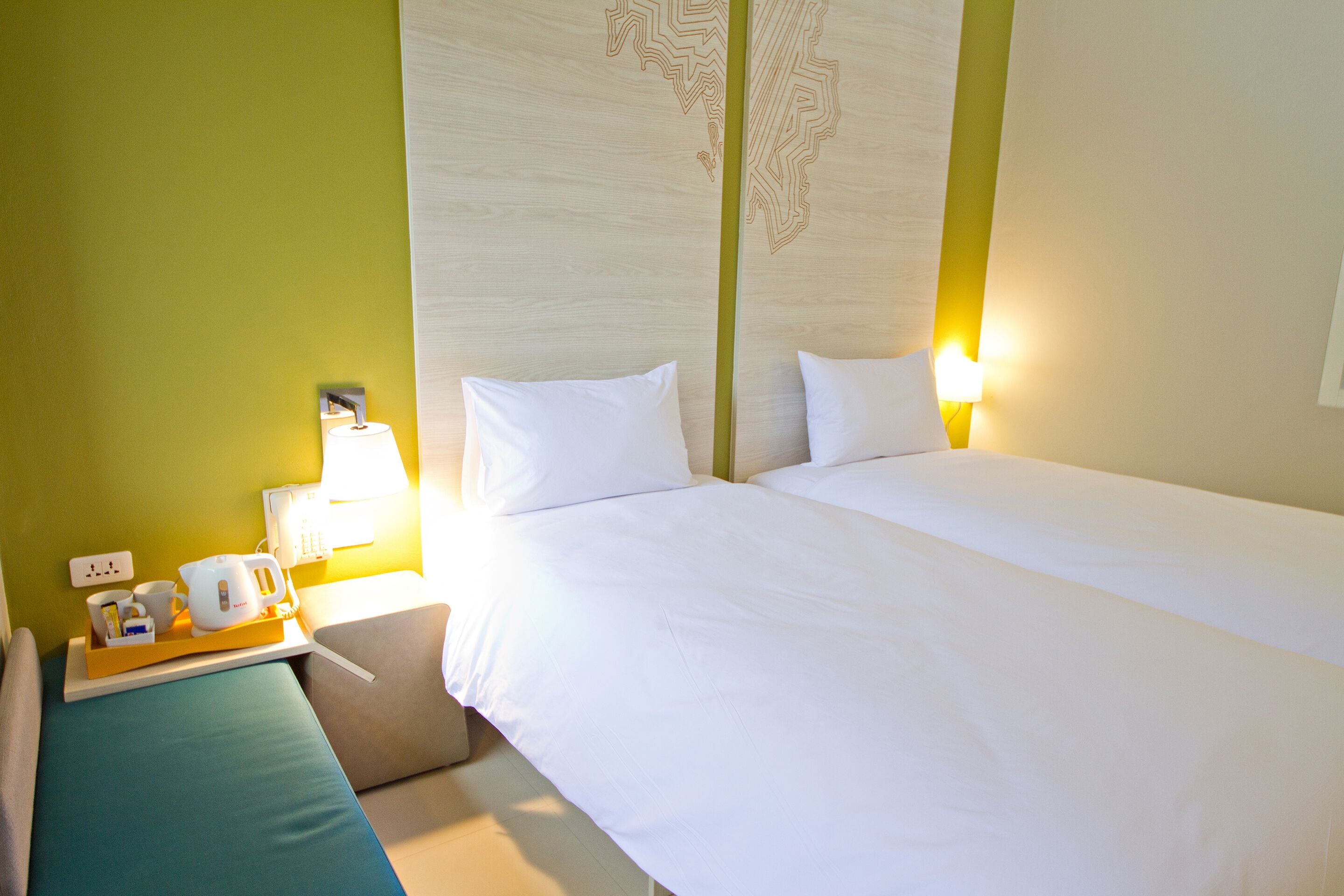 Habitación de hotel con cama, mesita de noche, lámpara y bandeja con tetera y tazas.