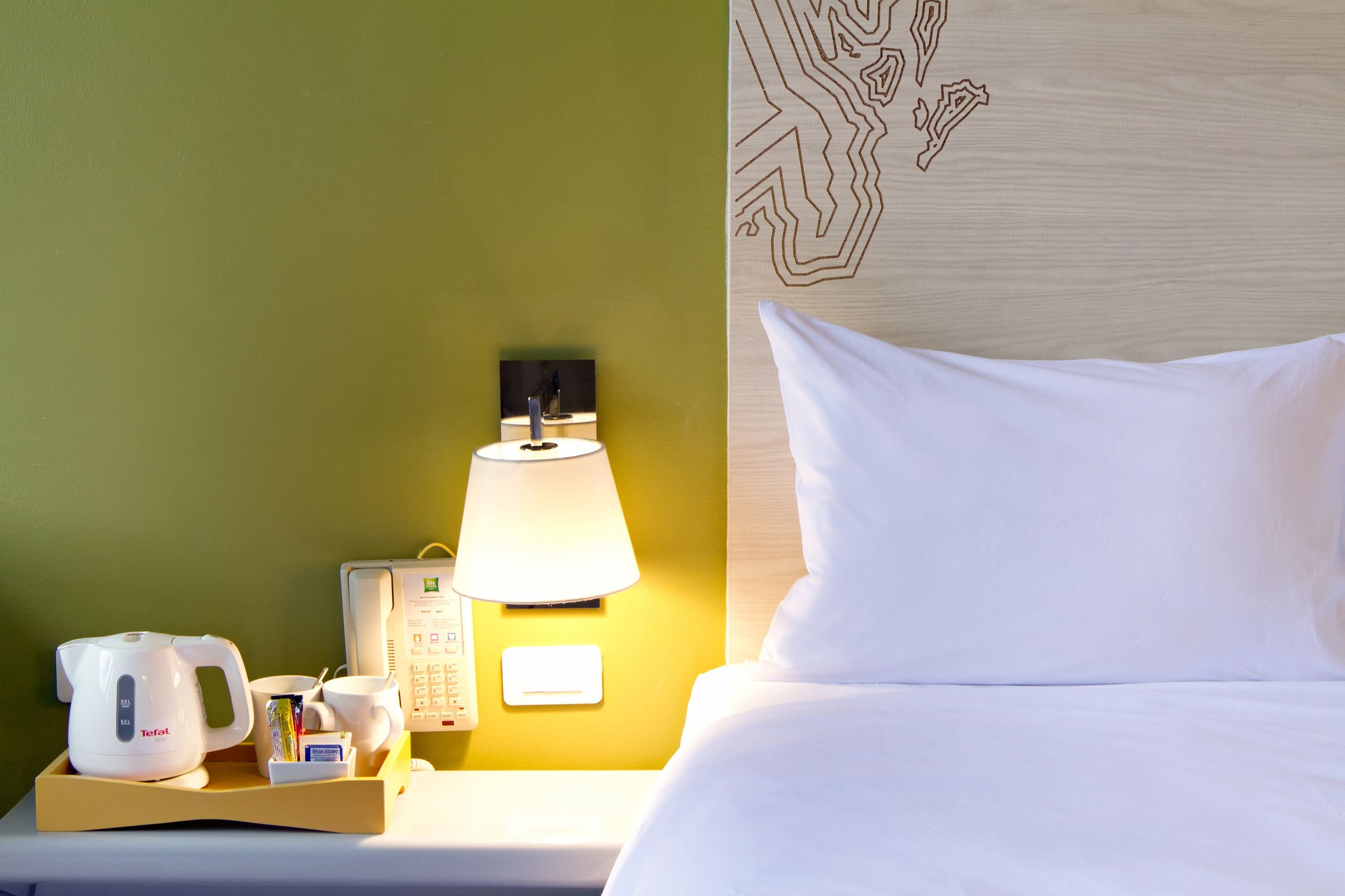 A hotel room with a bed, a nightstand with a lamp, a telephone, and a tray with a kettle, cups, and a mug.