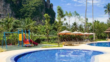 Relax by the pool surrounded by lush greenery and palm trees, with a children's play area nearby.