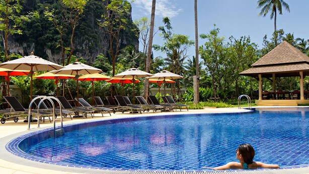 A swimming pool with lounge chairs and umbrellas, surrounded by palm trees and a mountain backdrop. - ibis Styles Krabi Ao Nang (Krabi)