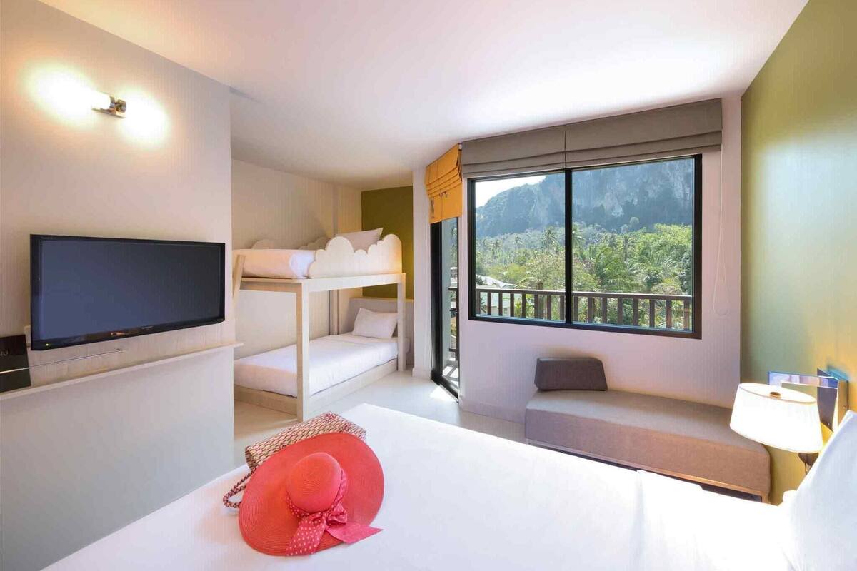 Une chambre d’hôtel comprenant un lit, une télévision, un lit superposé, une fenêtre donnant sur un espace verdoyant et un balcon.