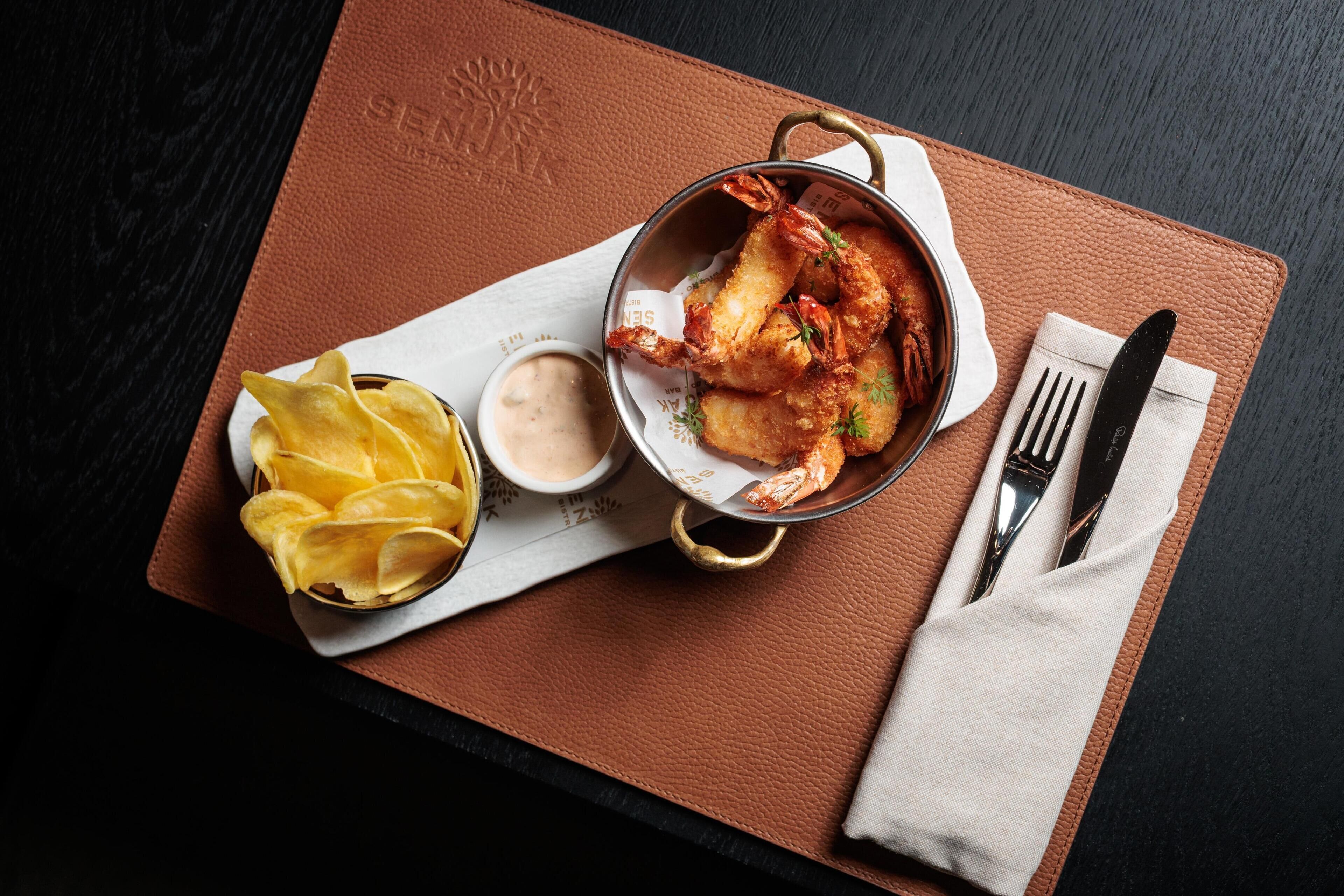 A meal of fried shrimp, chips, and dipping sauce on a brown leather table with a napkin and cutlery.