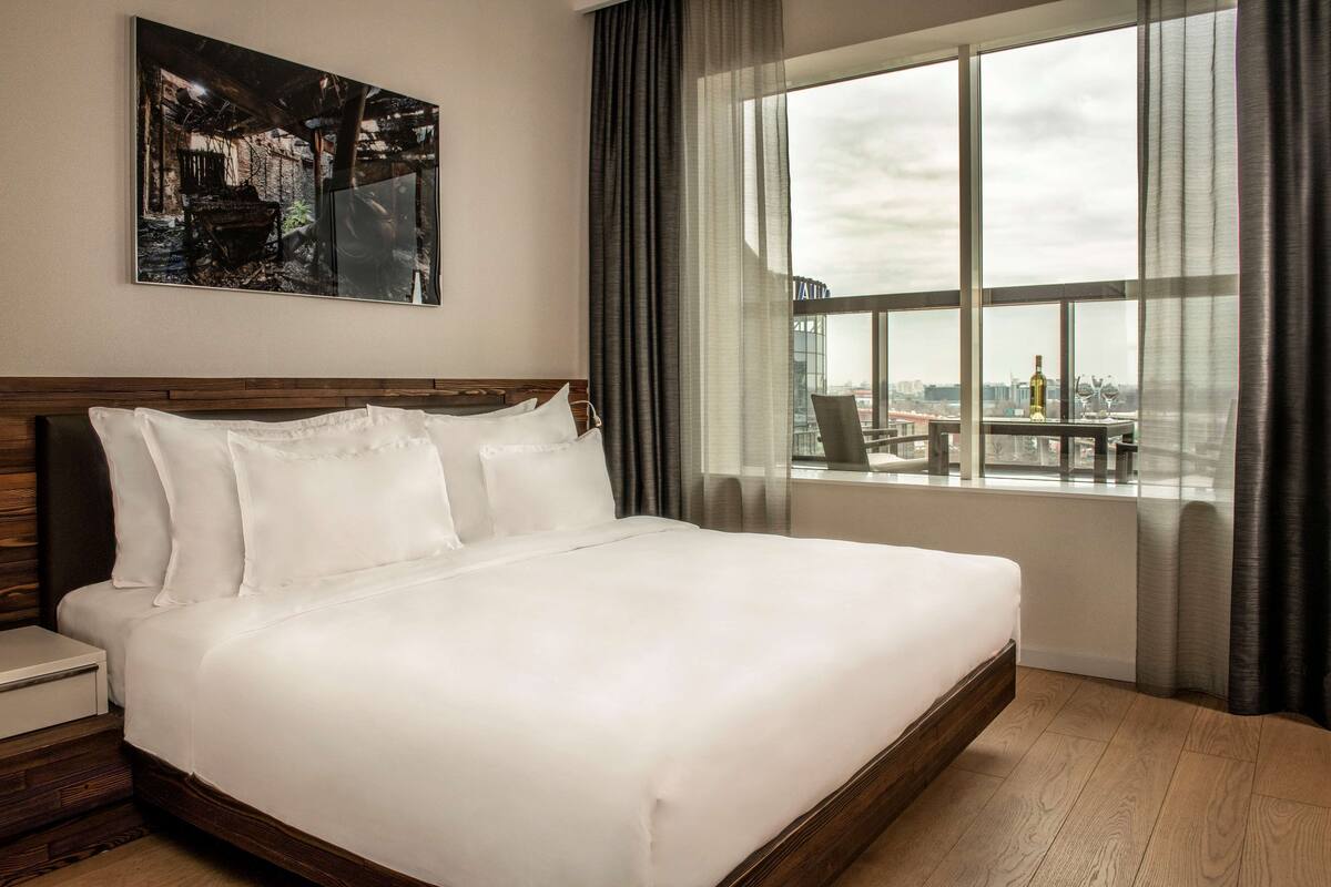 A large bed with white linens, a wooden headboard, and a nightstand with a drawer. A large window with curtains and a view of the city is visible.