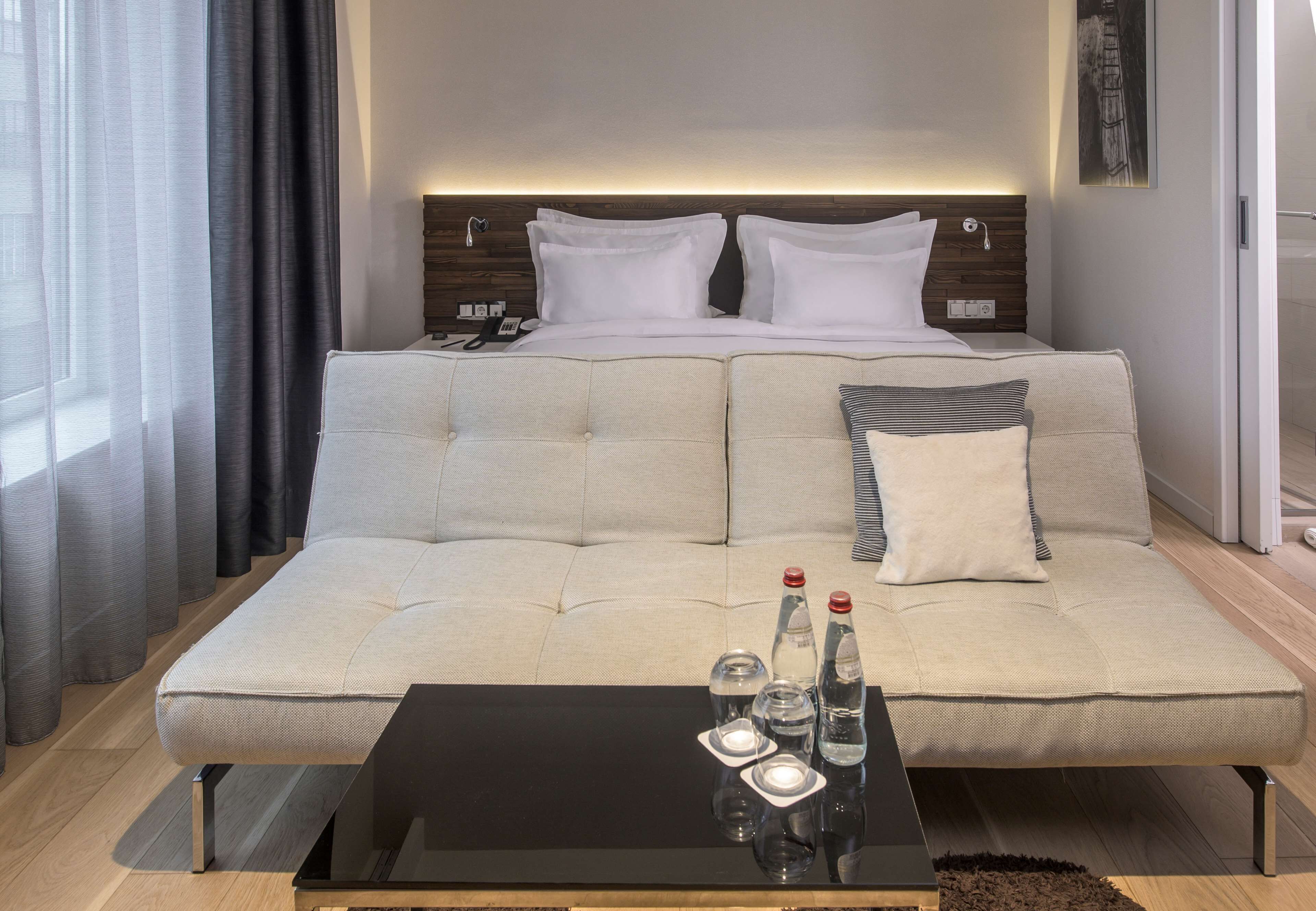 A modern hotel room with a large bed, a sofa, a glass coffee table, and a bedside table with a phone and a lamp.