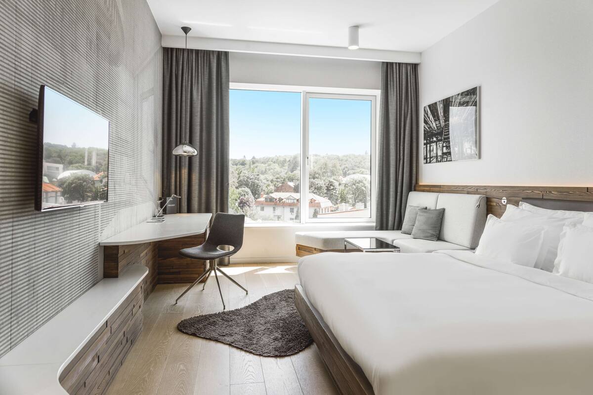 A modern hotel room with a large bed, a desk with a chair, a flat-screen TV, and a large window offering a view of the outdoors.