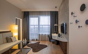 A modern hotel room with a sofa, a small table with glasses, a desk with a chair, a TV, and a large window with curtains.