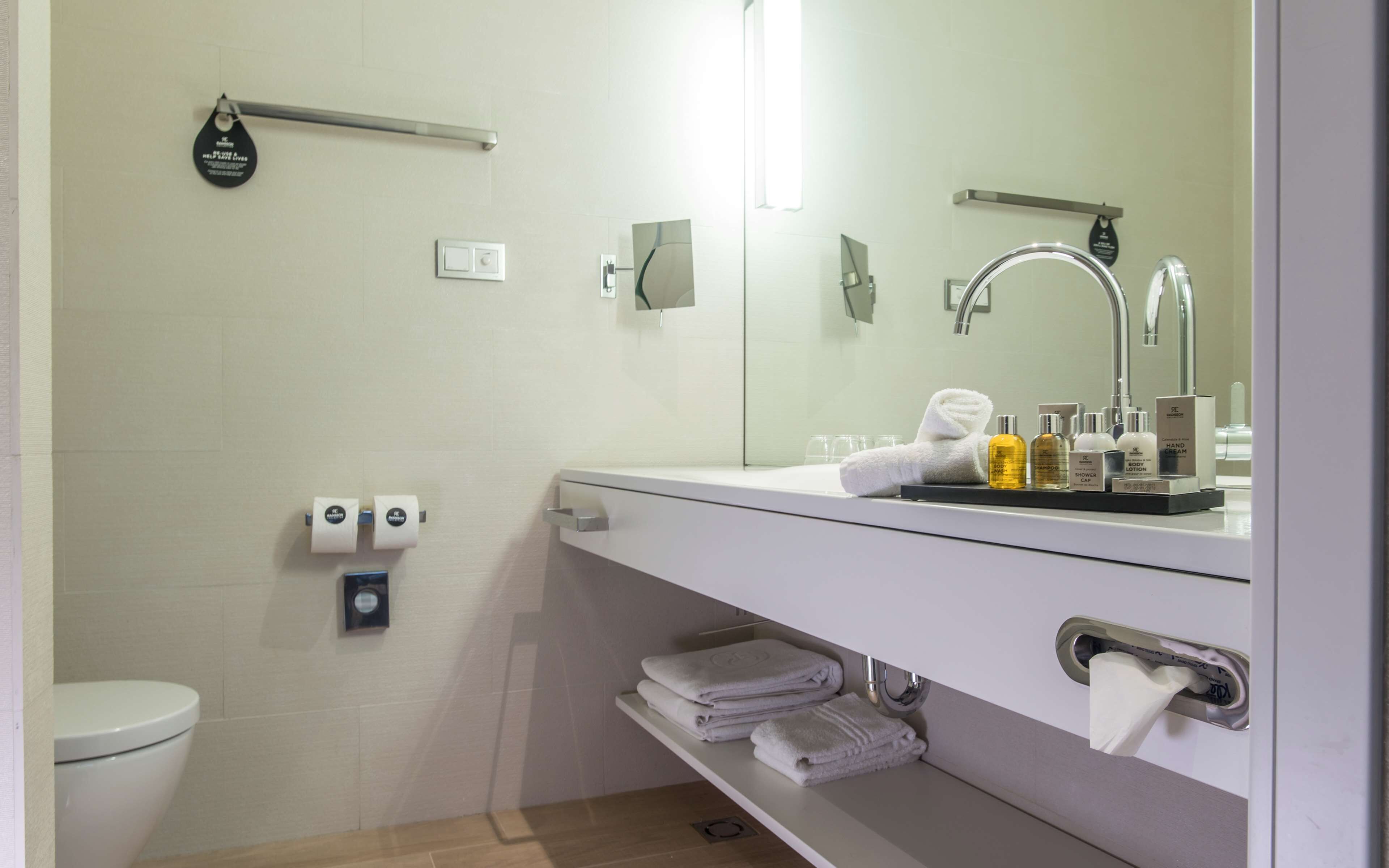 A modern bathroom with a white sink, mirror, and towel rack. There are toiletries on the sink counter, and a toilet with a paper towel dispenser.
