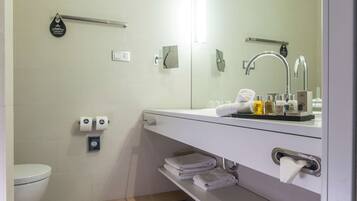A modern bathroom with a white sink, mirror, and towel rack. There are toiletries on the sink counter, and a toilet with a paper towel dispenser.