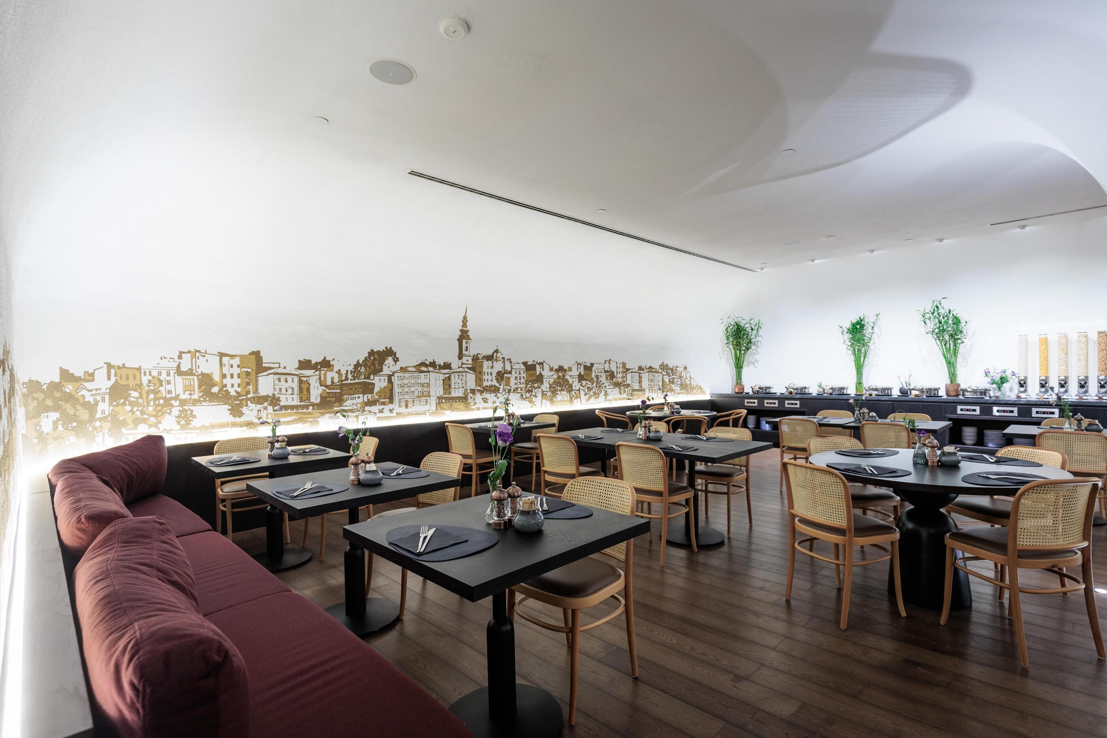 Un restaurant dont un mur est décoré d’une fresque représentant un paysage urbain, avec des tables et des chaises en bois, et un coin salon doté de sièges rembourrés.