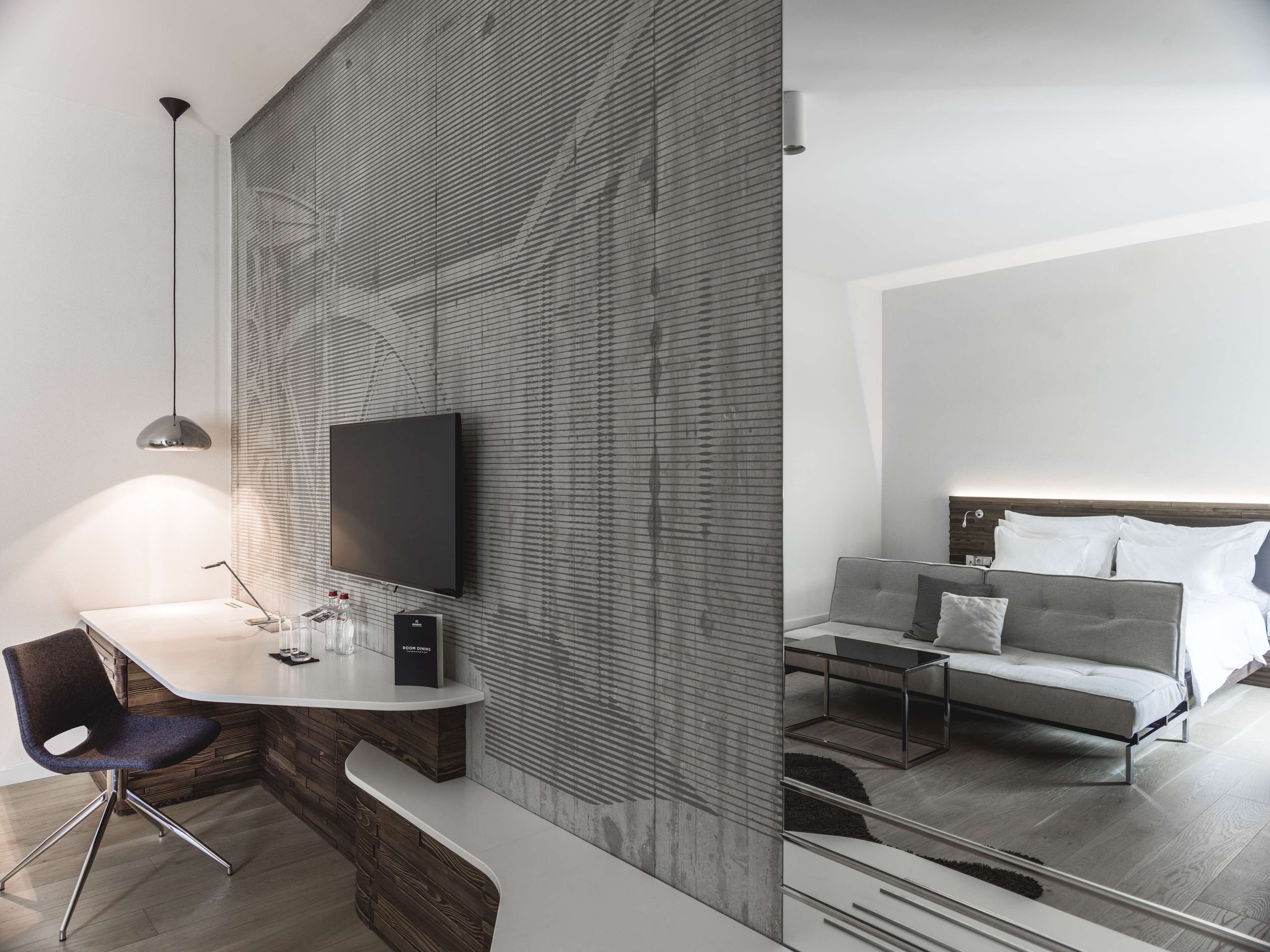 Una habitación de hotel moderna con una televisión de pantalla plana montada en una pared con textura, un escritorio con una silla y una cama sofá con cabecera.