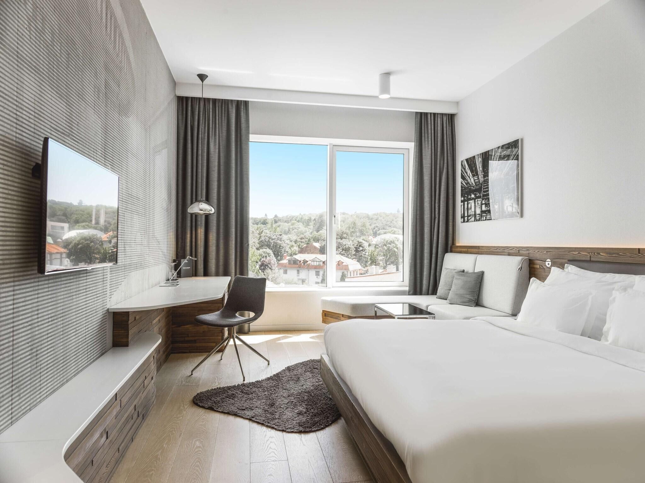 A modern hotel room with a bed, a desk, a chair, a TV, and a large window offering a view of the outside.