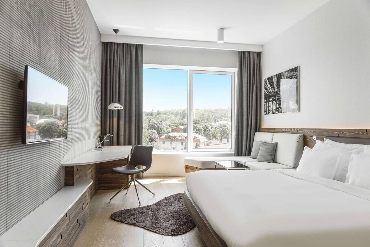 A modern hotel room with a bed, a desk, a chair, a TV, and a large window offering a view of the outside.