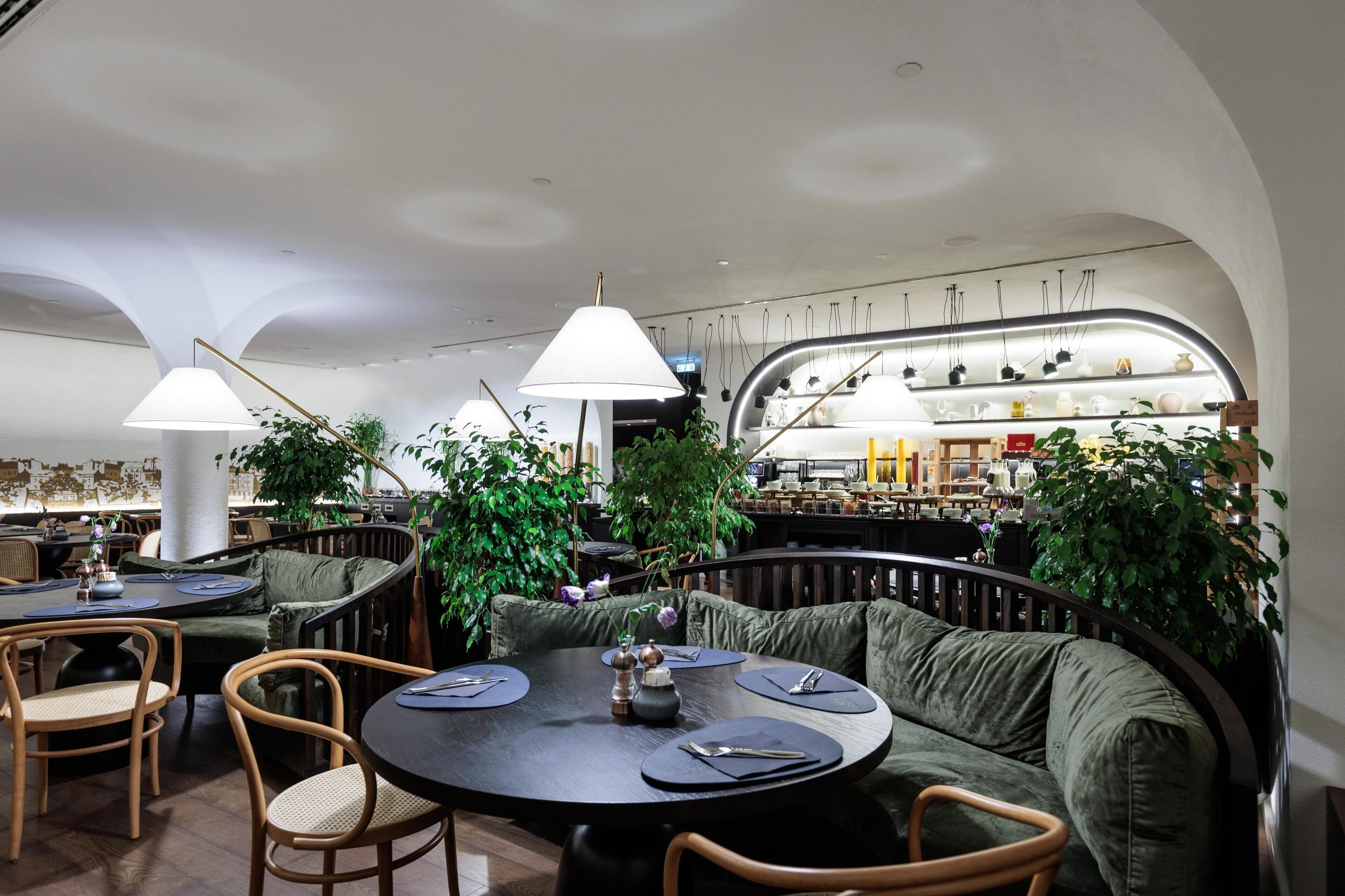 A restaurant with round tables, green cushioned seating, and hanging pendant lights.