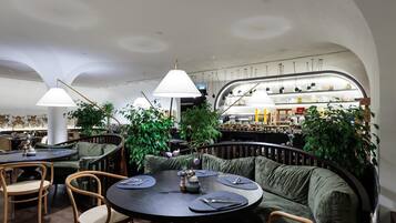 A restaurant with round tables, green cushioned seating, and hanging pendant lights.