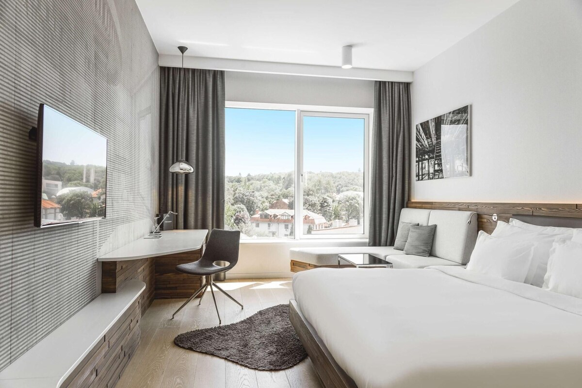 Una habitación de hotel moderna con cama, escritorio, silla, televisor y un ventanal con vistas al exterior.