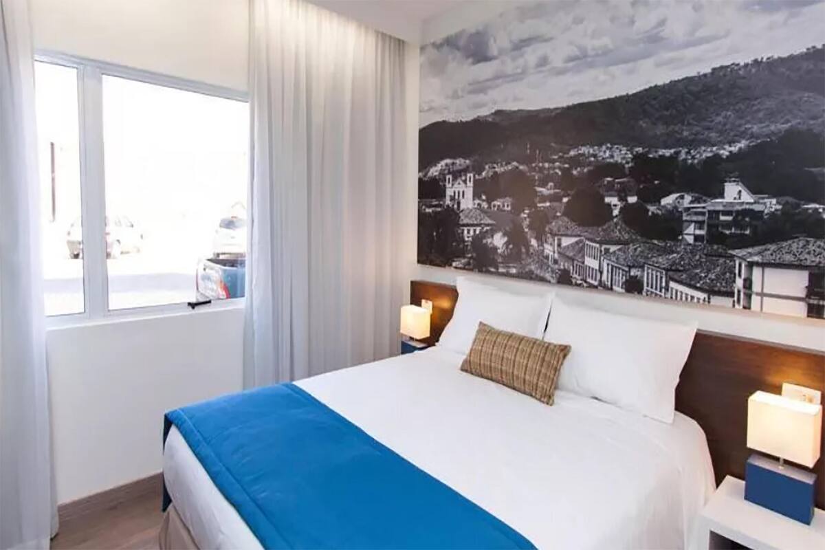 A hotel room with a large bed, a window with curtains, a nightstand with a lamp, and a wall-mounted painting of a town.