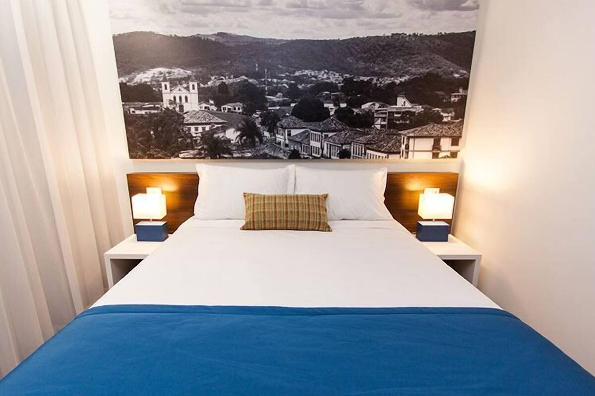 A hotel room with a bed, two bedside lamps, a plaid pillow, and a large landscape painting on the wall.