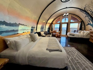 Ein gemütliches Schlafzimmer mit einem großen Bett, einem hölzernen Kopfteil und Blick ins Freie durch ein großes Fenster.