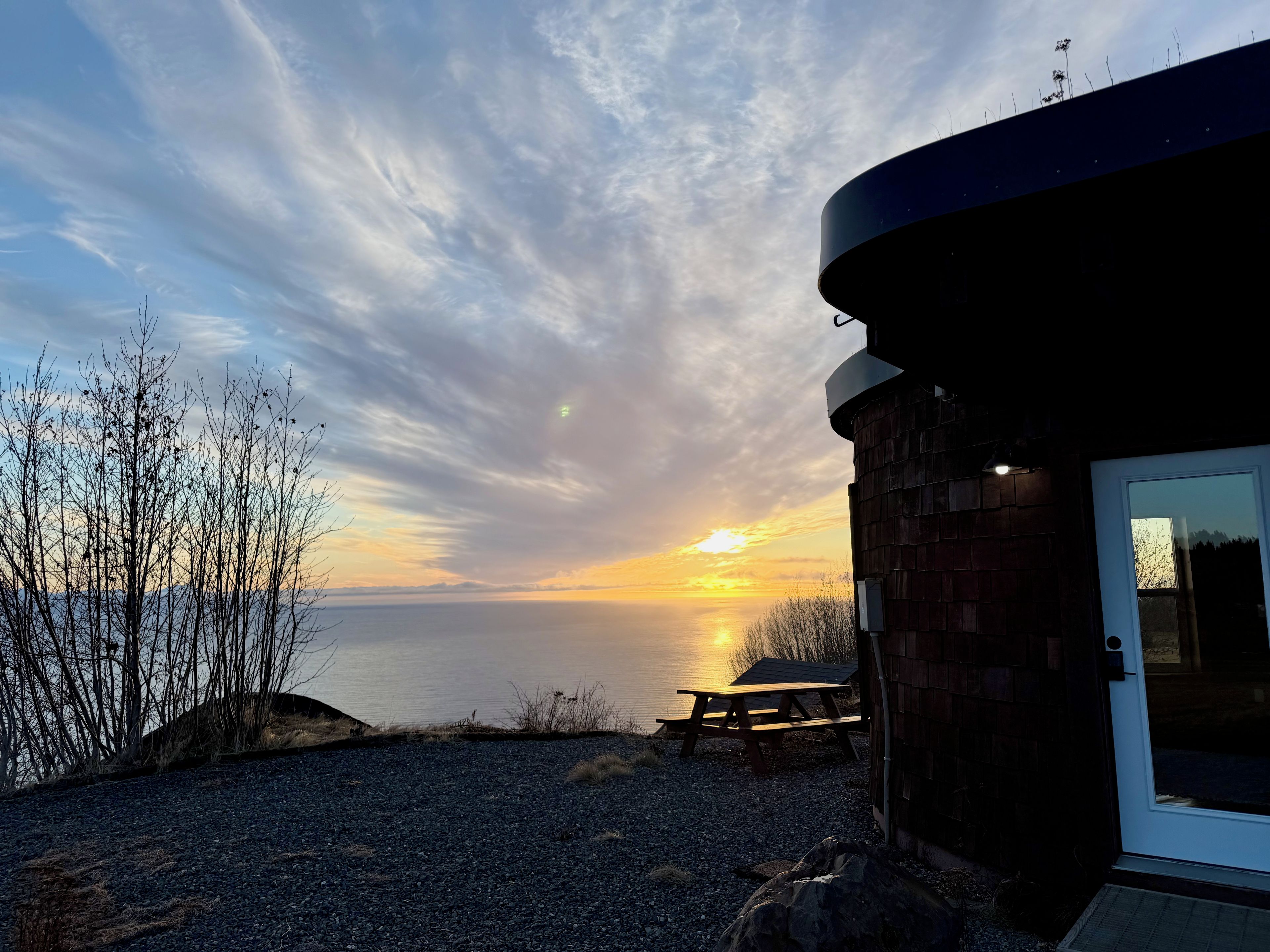 Un bâtiment avec une porte et une fenêtre, une table de pique-nique et une vue sur le coucher de soleil sur l’eau.