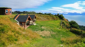 A coastal property with a grassy lawn, two wooden cabins, and a view of the sea and mountains. - Kenai Peninsula Suites (Homer)