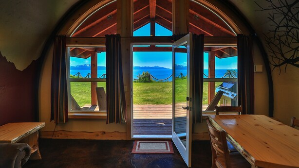 A room with a large arched window, wooden furniture, and a view of a grassy field and mountains. - Kenai Peninsula Suites (Homer)