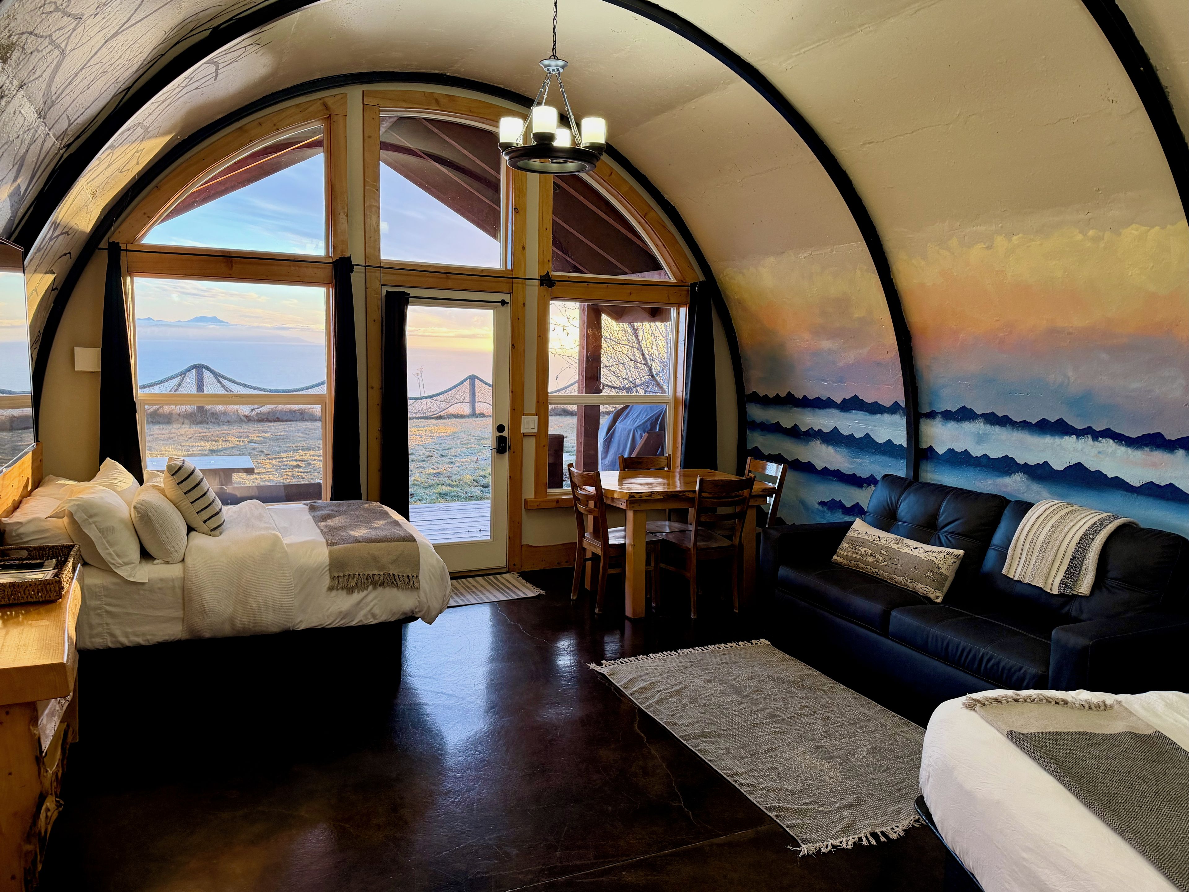 Interior acolhedor com cama, sofá, mesa de jantar e grandes janelas que oferecem vista para uma ponte e paisagem.