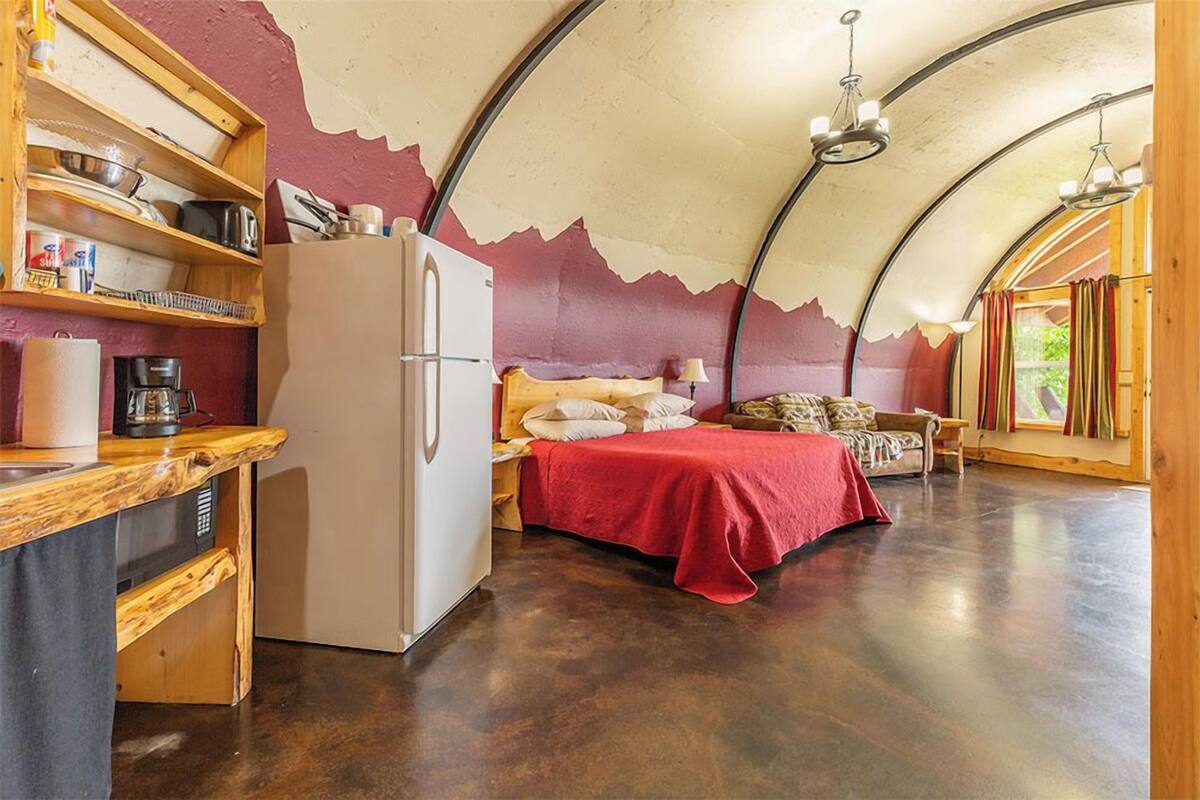 A compact room with a bed, a refrigerator, a wooden shelf with kitchen items, and a large window.