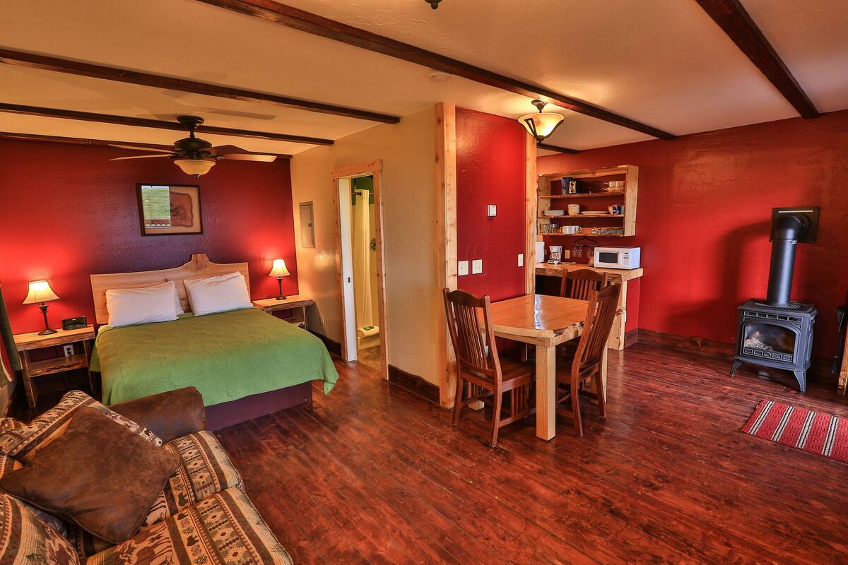 A cozy room with a bed, a dining table with chairs, a red wall, a ceiling fan, and a wood-burning stove.