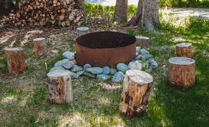 A circular fire pit surrounded by evenly spaced wooden stumps, set in a grassy area with trees and a pile of logs in the background. - Kenai Peninsula Suites (Homer)