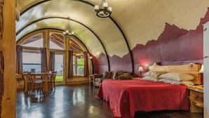 A spacious room with a curved ceiling, a large bed with a red bedspread, a wooden dining table, and a view through large windows. - Kenai Peninsula Suites (Homer)