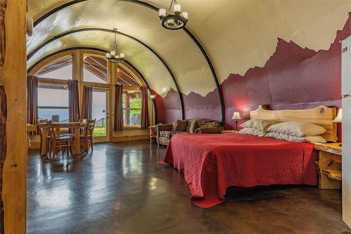 A spacious room with a curved ceiling, a large bed with a red bedspread, a wooden dining table, and a view through large windows.