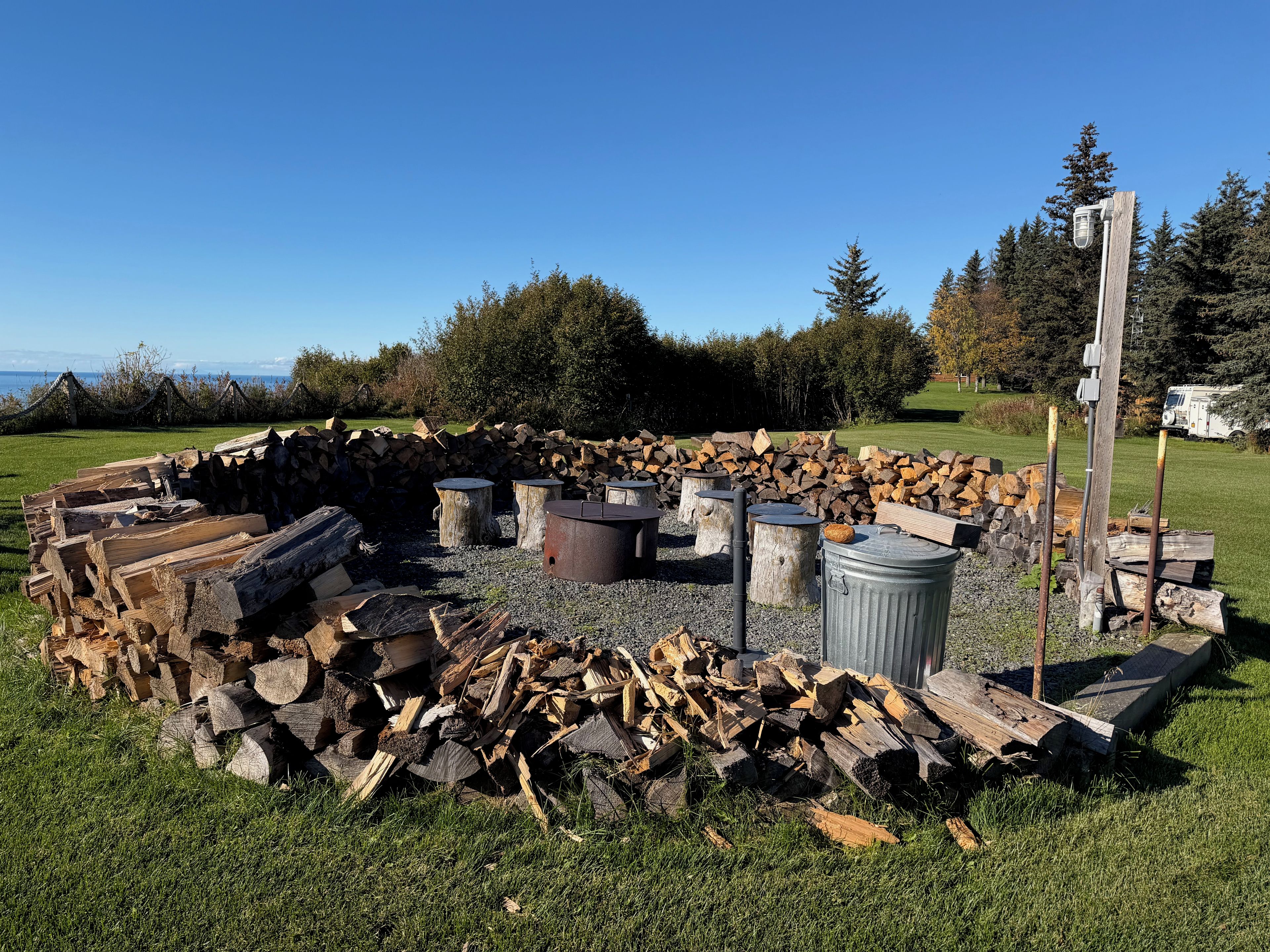 An outdoor fire pit area with logs, a metal container, and a wooden bench.