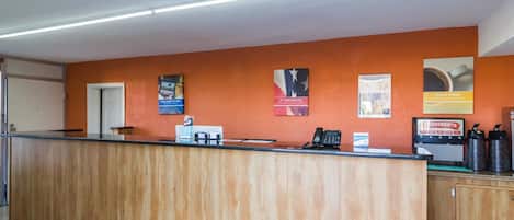 Un mostrador de recepción con encimera de madera, un teléfono y una máquina de café. Hay carteles en la pared y una puerta a la izquierda.