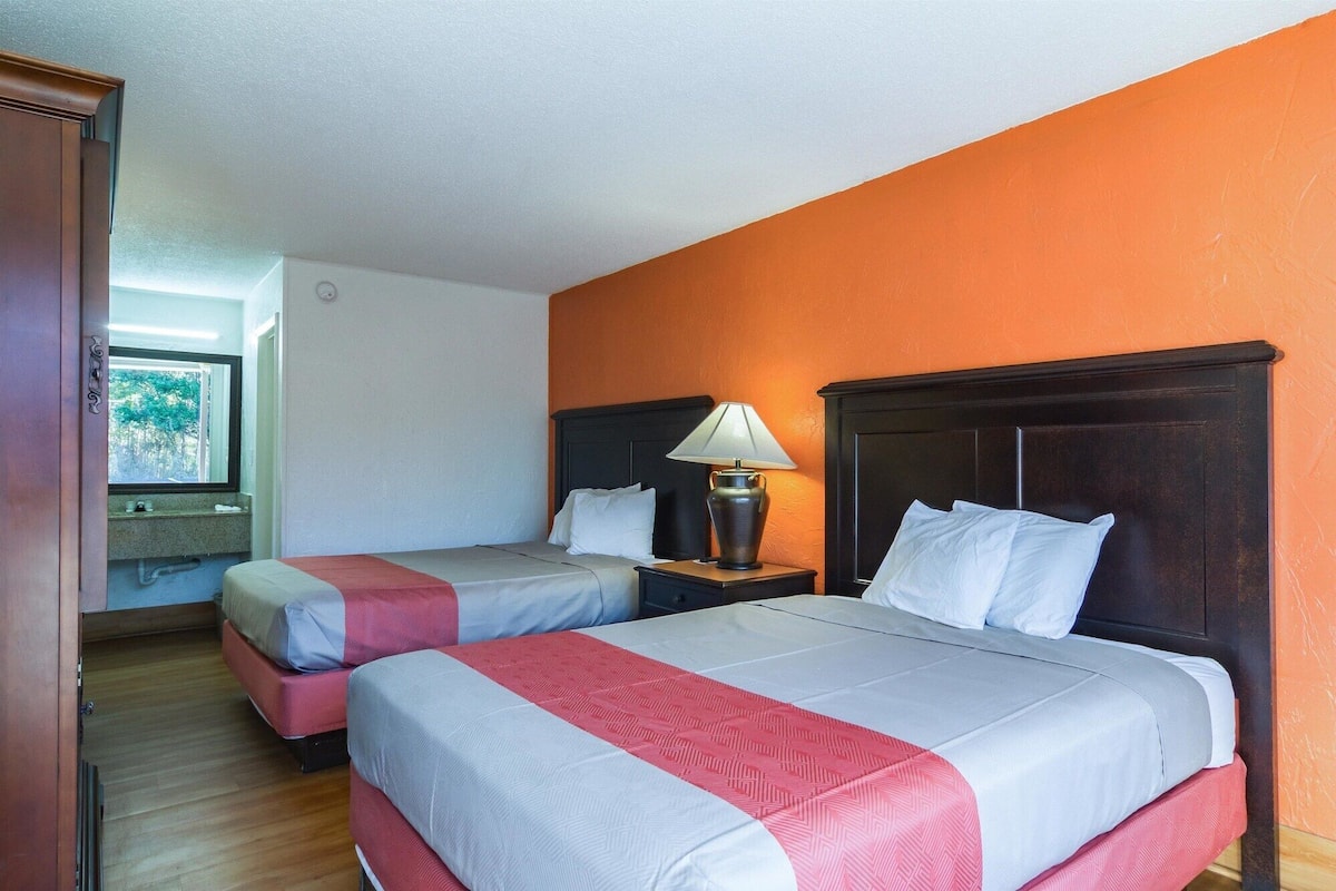 A hotel room with two beds, a nightstand with a lamp, and a window with a view of trees.