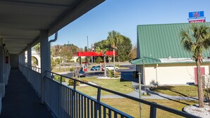A covered walkway with a view of a gas station, a building with a green roof, and a palm tree. - Motel 6 Townsend, GA (Townsend)