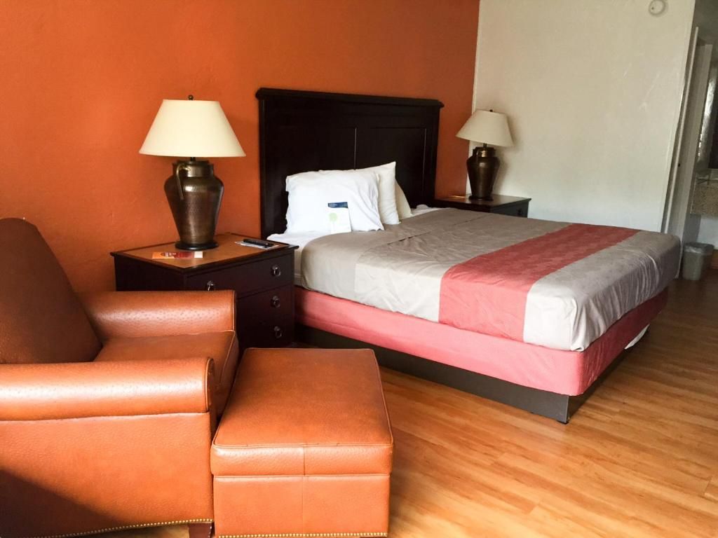 Quarto de hotel com cama, mesa de cabeceira, luminária, cadeira e piso de madeira.