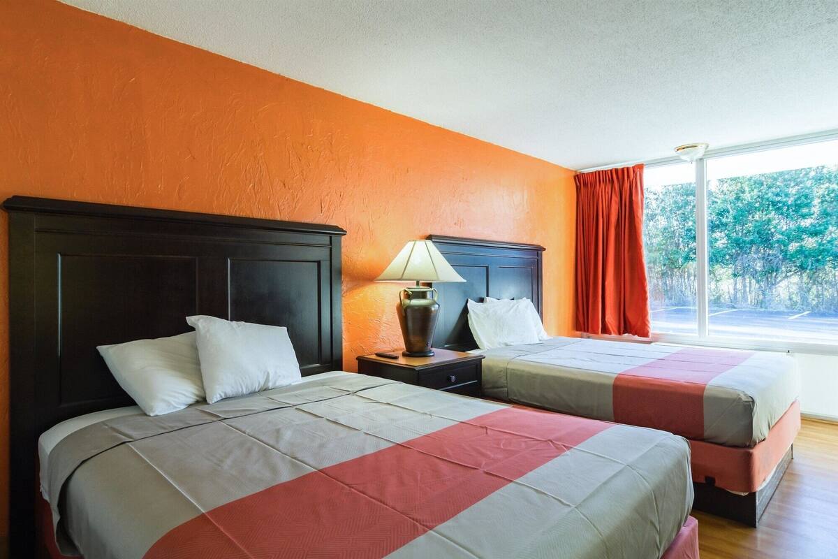 A hotel room with two beds, a nightstand with a lamp, and a window with a view of trees.