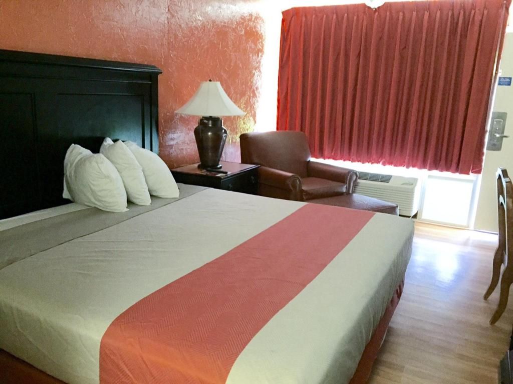 Una habitación de hotel con cama, mesita de noche, lámpara, silla y ventana con cortinas.