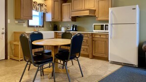 Una cocina compacta con armarios de madera, un refrigerador blanco, un microondas, una mesa de comedor con cuatro sillas y una ventana con cortinas.