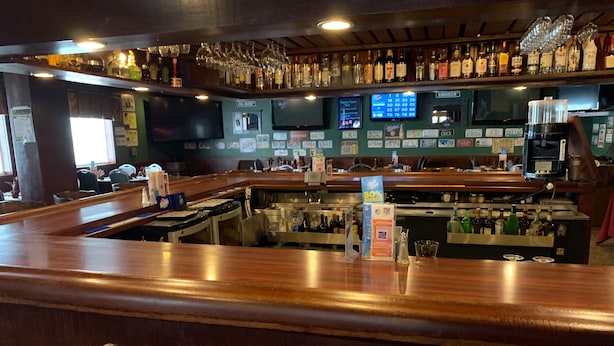 Zona de bar con mostrador de madera, taburetes y una variedad de botellas y vasos en estanterías.
