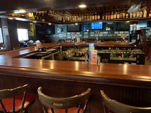 Zona de bar con mostrador de madera, taburetes y una variedad de botellas y vasos en estanterías.