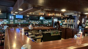 Una zona de bar con mostrador de madera, estantes con botellas y una pantalla digital mostrando resultados.
