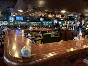 A bar area with a wooden counter, shelves with bottles, and a digital screen displaying scores. - Fred's Inn (Norwich)