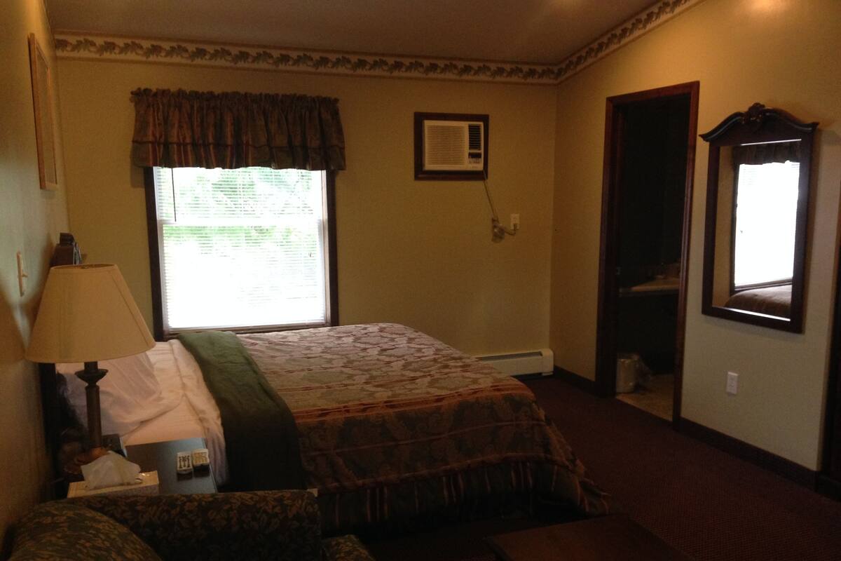 Une chambre d’hôtel comprenant un lit, une chaise, une lampe, une fenêtre avec des rideaux, un climatiseur fixé au mur et un miroir.