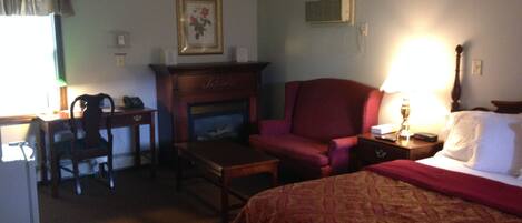 A hotel room with a bed, a desk, a chair, a fireplace, and a lamp.