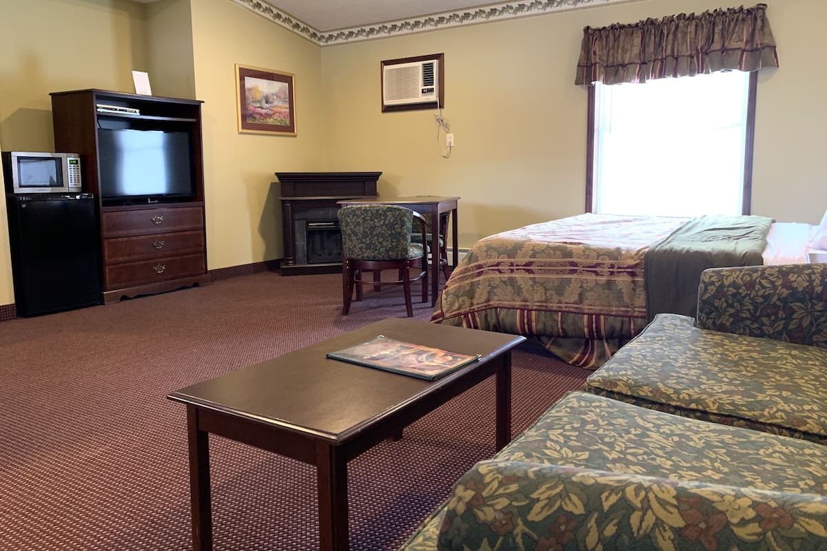 Une chambre d’hôtel avec un lit, deux fauteuils, une petite table, un téléviseur, une cheminée et un tableau au mur.