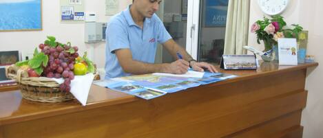 A man at a wooden reception desk, signing documents. There is a basket of fruit, a clock, and a framed picture on the wall behind him.