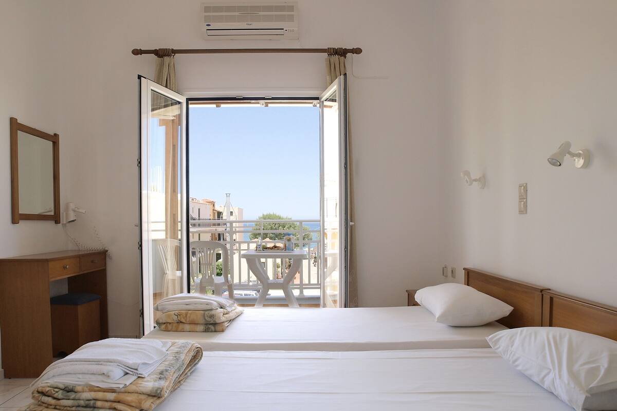 Ein Hotelzimmer mit Bett, Schreibtisch, Stuhl und einem Balkon mit Blick auf Gebäude und das Meer.